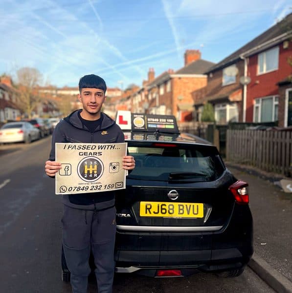 A smiling learner holds a Gears pass board beside a black Nissan Micra on a terraced street under a blue sky
