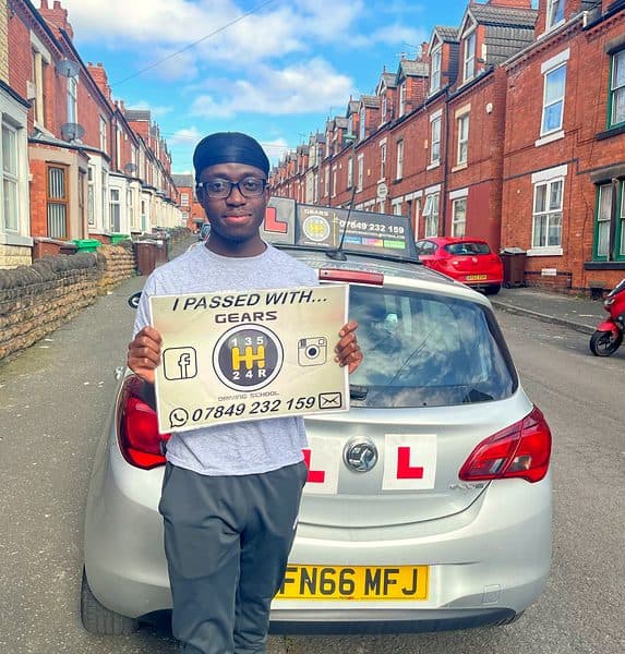 A learner in glasses holds a Gears pass board in front of a silver Vauxhall Corsa on a red-brick terraced street