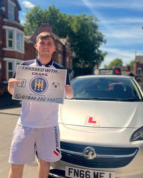 A smiling learner in a polo shirt holds a Gears pass board in front of a silver Vauxhall Corsa on a sunny day