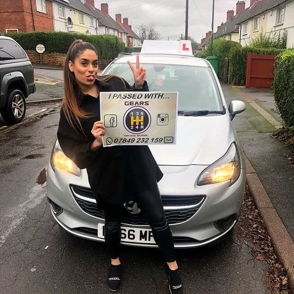 A learner makes a peace sign while holding a Gears pass board in front of a silver Vauxhall Corsa