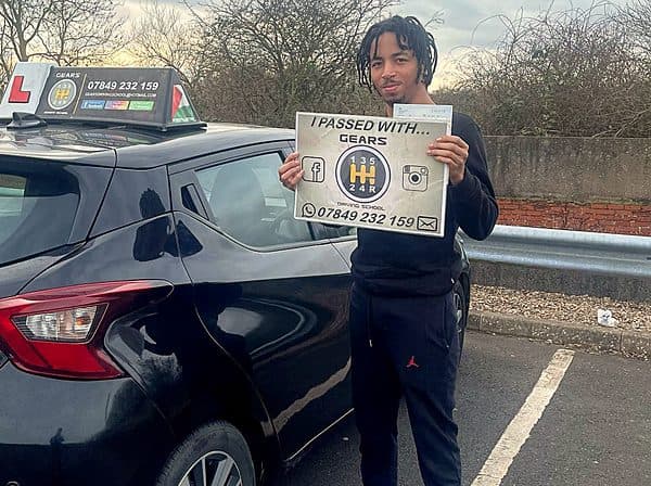 A learner holds a Gears pass board beside a black Nissan Micra in a car park