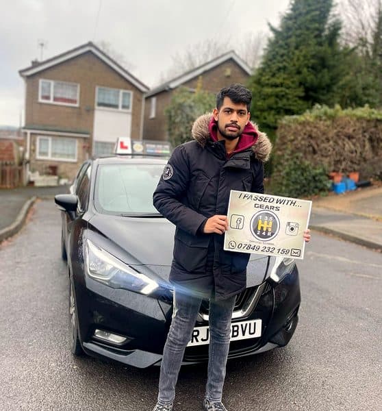 A learner in a fur-hooded parka holds a Gears pass board beside a black Nissan Micra