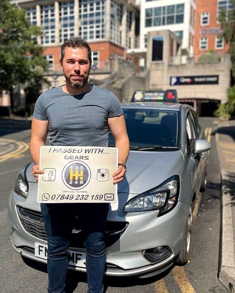 A learner in a grey T-shirt holds a Gears pass board in front of a silver Vauxhall Corsa in the city centre