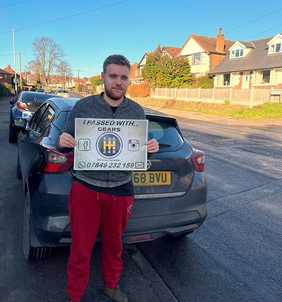 A learner in red trousers holds a Gears pass board beside a black Nissan Micra on a bright winter street