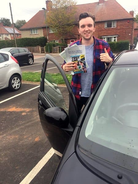 A grinning learner gives a thumbs up with a pass certificate beside a black Toyota Aygo with its door open