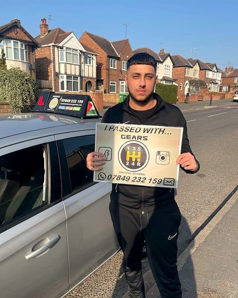 A learner holds a Gears pass board beside a silver Vauxhall Corsa on a sunny suburban street