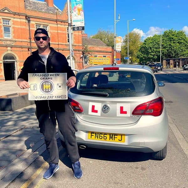 A learner in sunglasses holds a Gears pass board behind a silver Vauxhall Corsa on a sunny high street