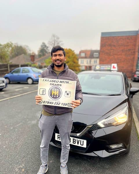 A smiling learner holds a Gears pass board beside a black Nissan Micra on an autumn street