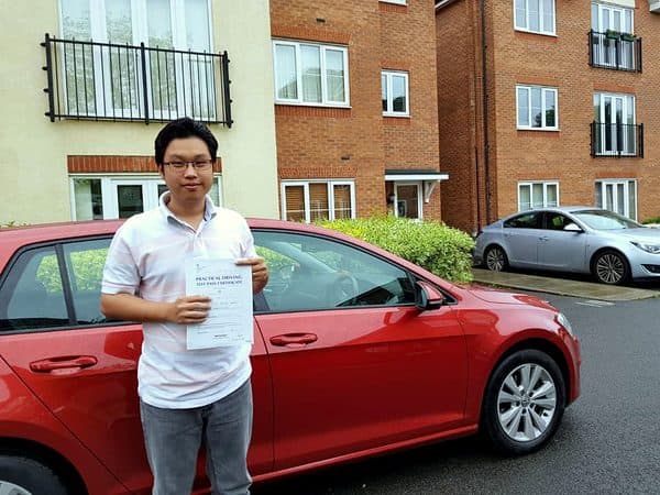 A learner in glasses holds a pass certificate in front of a red hatchback outside a block of flats