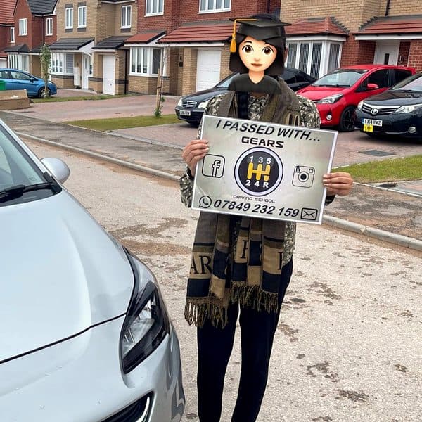 A learner in a long scarf, face covered by a graduate emoji, holds a Gears pass board beside a silver Vauxhall Corsa