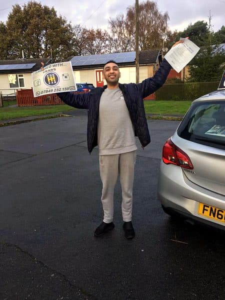A learner throws their arms in the air with a pass certificate and a Gears pass board beside a silver Vauxhall Corsa