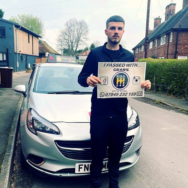 A learner in a black jacket holds a Gears pass board in front of a silver Vauxhall Corsa on a residential street