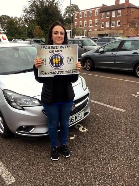 A smiling learner holds a Gears pass board in front of a silver Vauxhall Corsa in a car park
