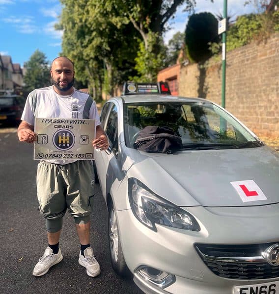 A smiling bearded learner holds a Gears pass board beside a silver Vauxhall Corsa on a leafy street