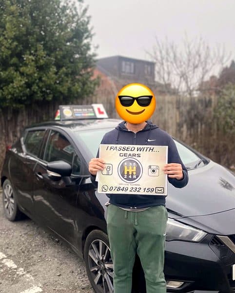 A learner, face covered by a sunglasses emoji, holds a Gears pass board beside a black Nissan Micra
