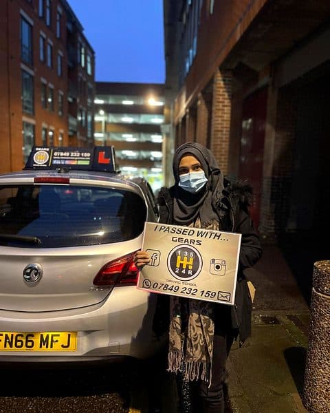 A learner in a headscarf and face mask holds a Gears pass board beside a silver Vauxhall Corsa on an evening street