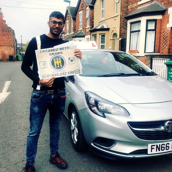 A learner in glasses holds a Gears pass board beside a silver Vauxhall Corsa
