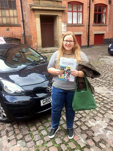 A smiling learner holds a pass certificate beside a black Toyota Aygo on a cobbled street