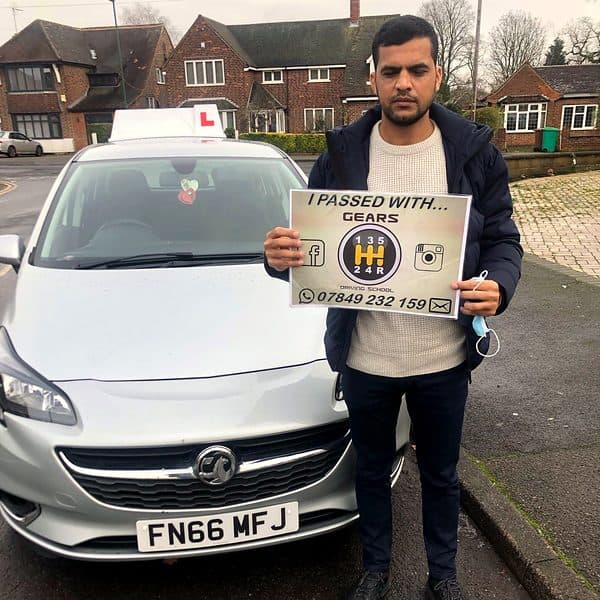 A learner holds a Gears pass board in front of a silver Vauxhall Corsa on a residential street