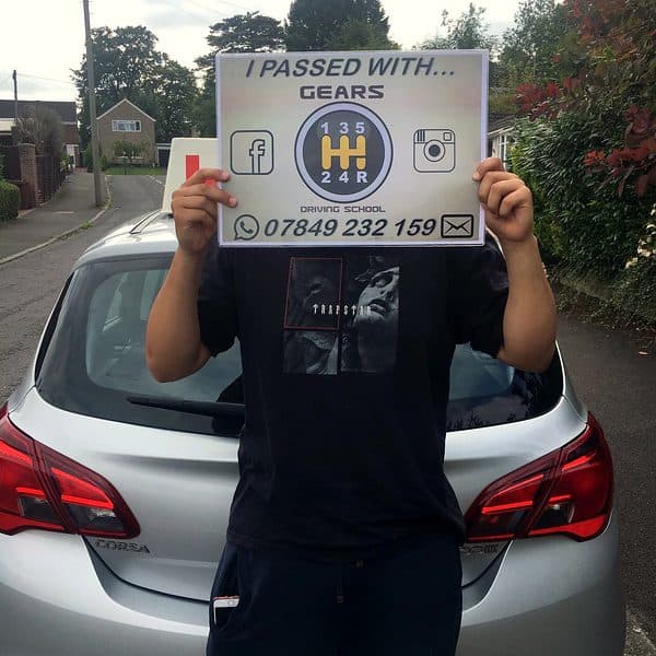 A learner holds a Gears pass board up over their face behind a silver Vauxhall Corsa
