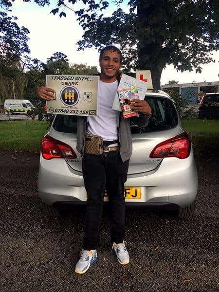 A smiling learner holds a Gears pass board and a theory leaflet behind a silver Vauxhall Corsa