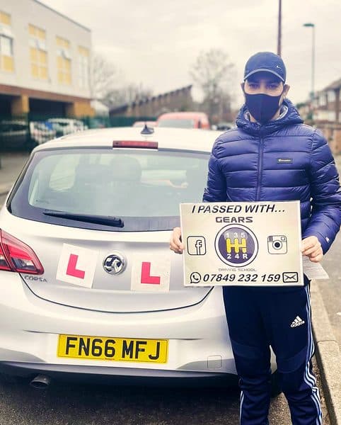 A learner in a cap, face mask and padded jacket holds a Gears pass board beside a silver Vauxhall Corsa