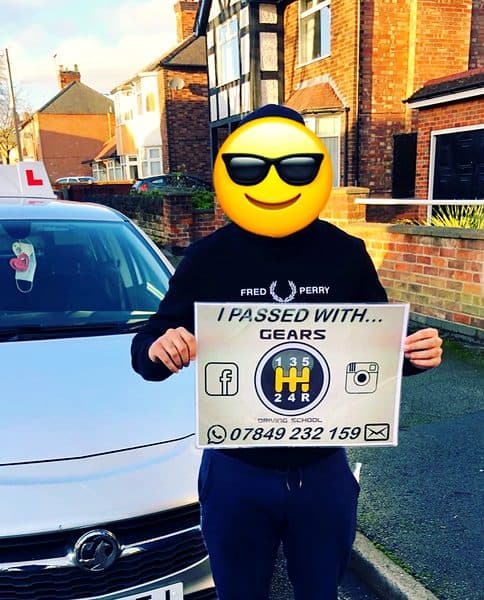 A learner, face covered by a sunglasses emoji, holds a Gears pass board in front of a silver Vauxhall Corsa