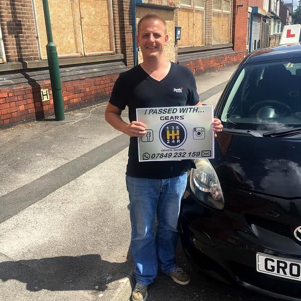 A smiling learner holds a Gears pass board beside a black Toyota Aygo