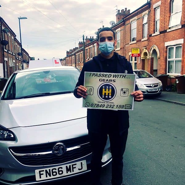 A learner in a face mask holds a Gears pass board in front of a silver Vauxhall Corsa on a terraced street