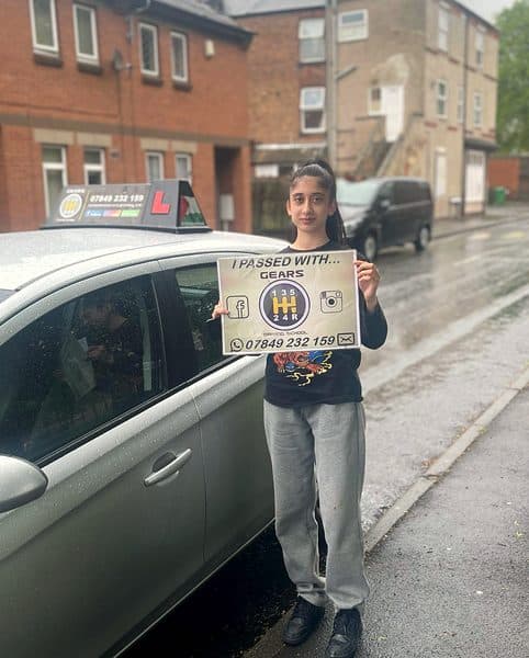 A learner holds a Gears pass board beside a silver Vauxhall Corsa on a wet street