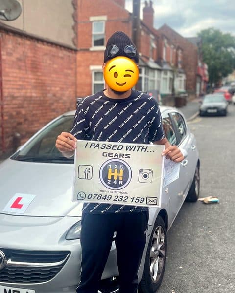 A learner, face covered by a winking emoji, holds a Gears pass board in front of a silver Vauxhall Corsa