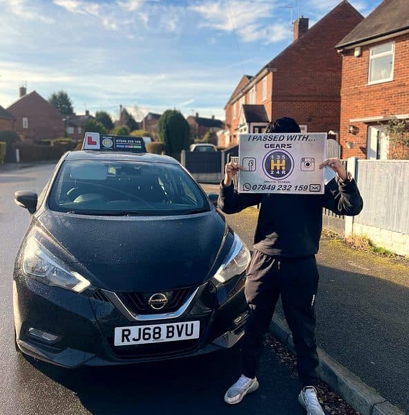 A learner raises a Gears pass board in front of their face beside a black Nissan Micra