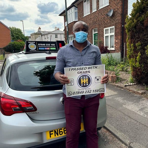 A learner in a face mask holds a Gears pass board behind a silver Vauxhall Corsa