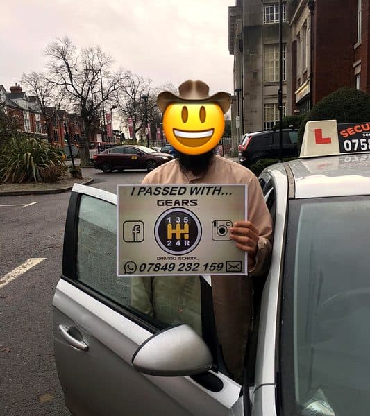 A learner, face covered by an emoji, holds a Gears pass board at the open door of a silver Vauxhall Corsa