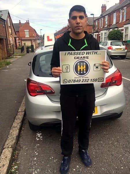 A learner holds a Gears pass board behind a silver Vauxhall Corsa on a terraced street