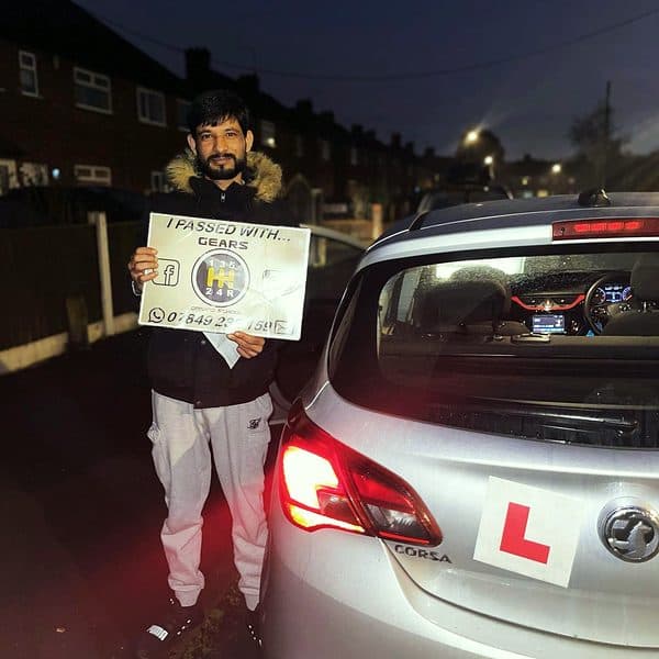At night, a learner holds a Gears pass board behind a silver Vauxhall Corsa with its lights on