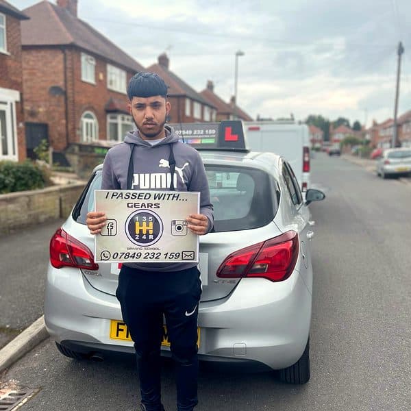A learner holds a Gears pass board behind a silver Vauxhall Corsa on a suburban street