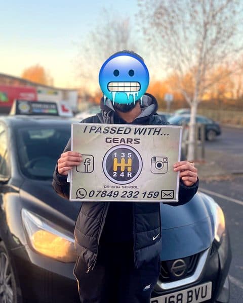 A learner, face covered by an emoji, holds a Gears pass board in front of a black Nissan Micra