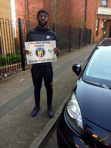A learner holds a Gears pass board beside a black Toyota Aygo on a pavement lined with railings