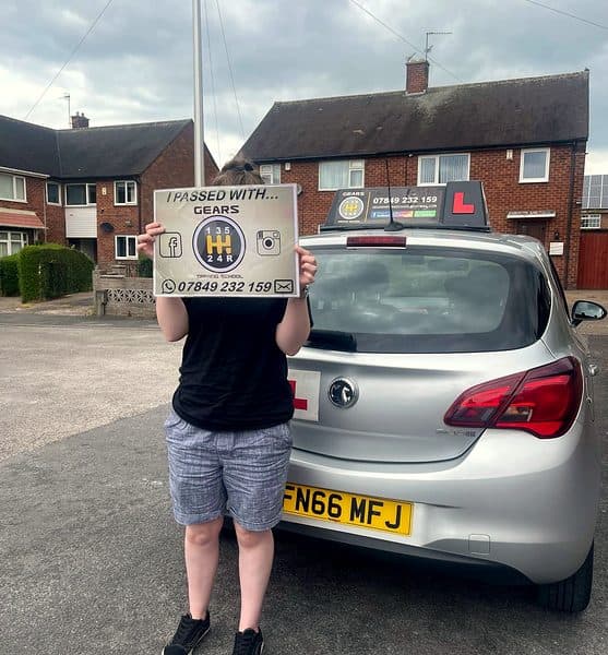 A learner hides their face behind a Gears pass board, standing beside a silver Vauxhall Corsa