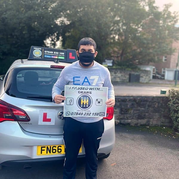 A learner in a face mask holds a Gears pass board behind a silver Vauxhall Corsa