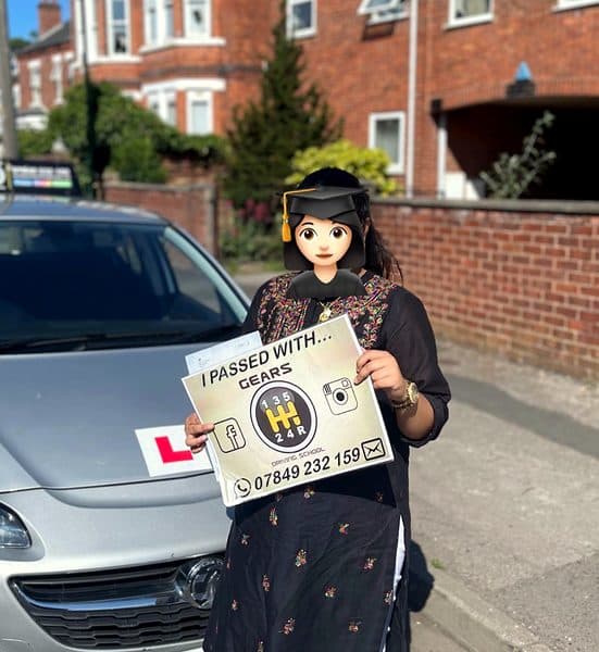 A learner, face covered by a graduate emoji, holds a Gears pass board beside a silver Vauxhall Corsa on a sunny day