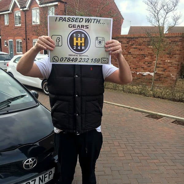 A learner holds a Gears pass board over their face beside a black Toyota Aygo