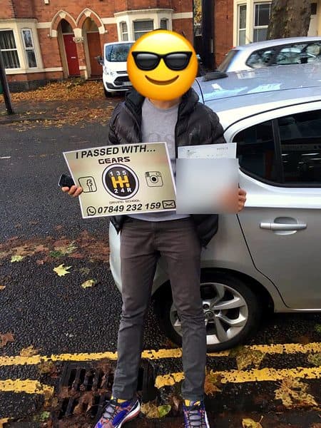 A learner, face covered by a sunglasses emoji, holds a Gears pass board and a pass certificate beside a silver Vauxhall Corsa on an autumn street