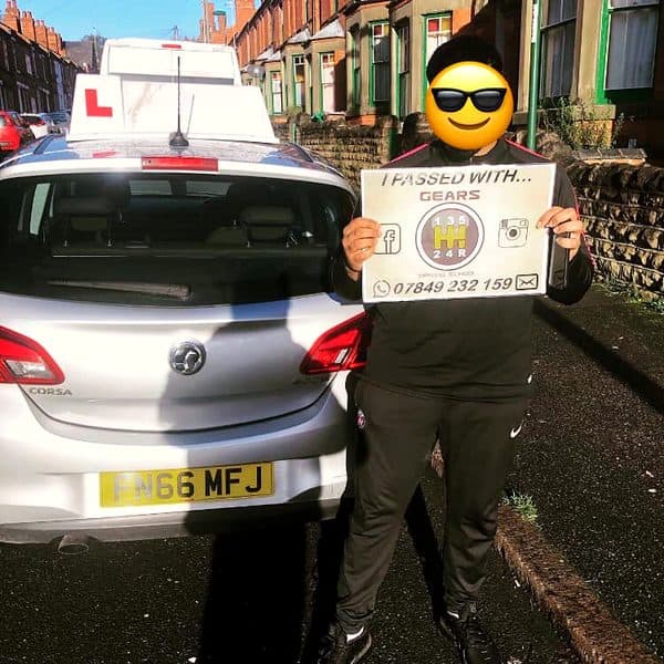 A learner, face covered by a sunglasses emoji, holds a Gears pass board behind a silver Vauxhall Corsa