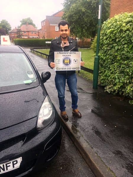 A learner holds a Gears pass board beside a black Toyota Aygo on a rainy street