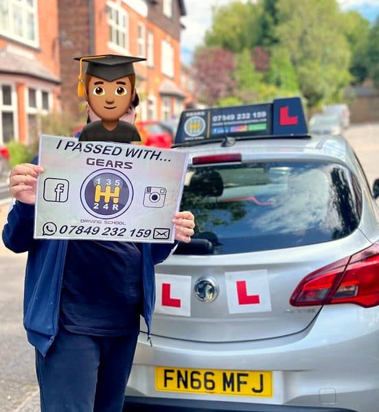 A learner, face covered by a graduate emoji, holds a Gears pass board behind a silver Vauxhall Corsa