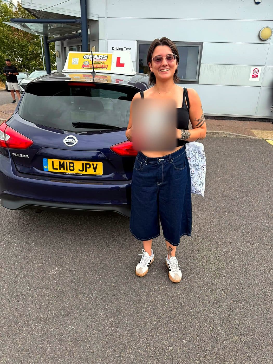 A smiling learner in sunglasses holds a pass certificate behind a blue Nissan Pulsar outside the driving test centre