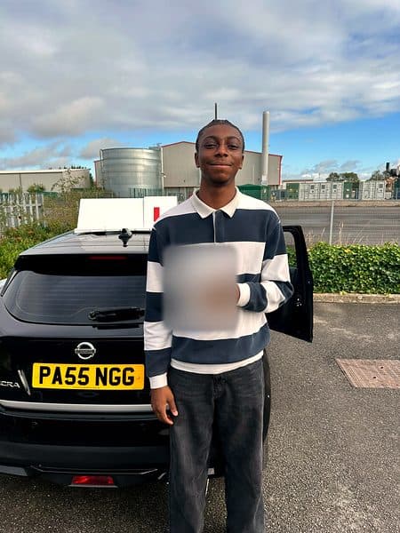 A smiling learner in a striped rugby shirt holds a pass certificate beside a black Nissan Micra