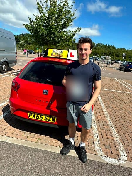A learner in a navy T-shirt holds a pass certificate beside a red SEAT Ibiza on a sunny day